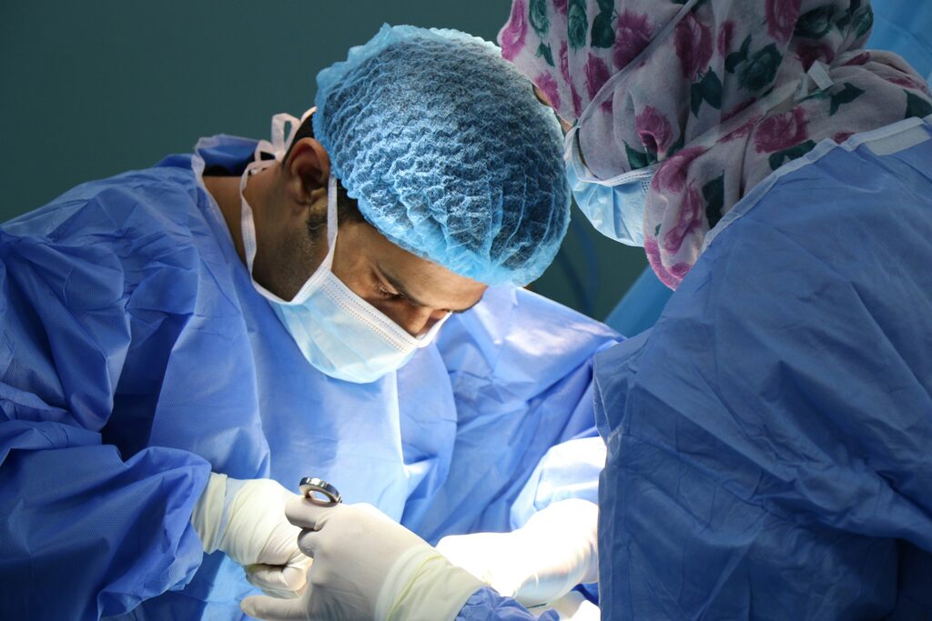 Médico usando toca e máscara, vestindo traje azul durante um procedimento cirúrgico.