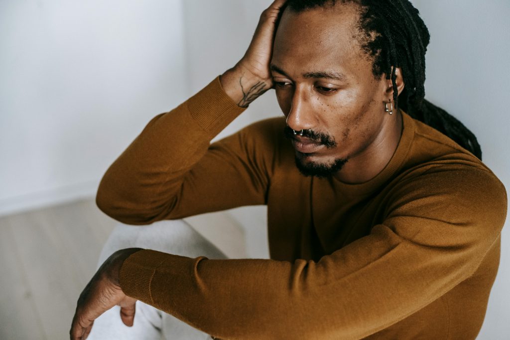 Homem negro de dreads e piercings levando a mão direita a cabeça em estado pensativo.
