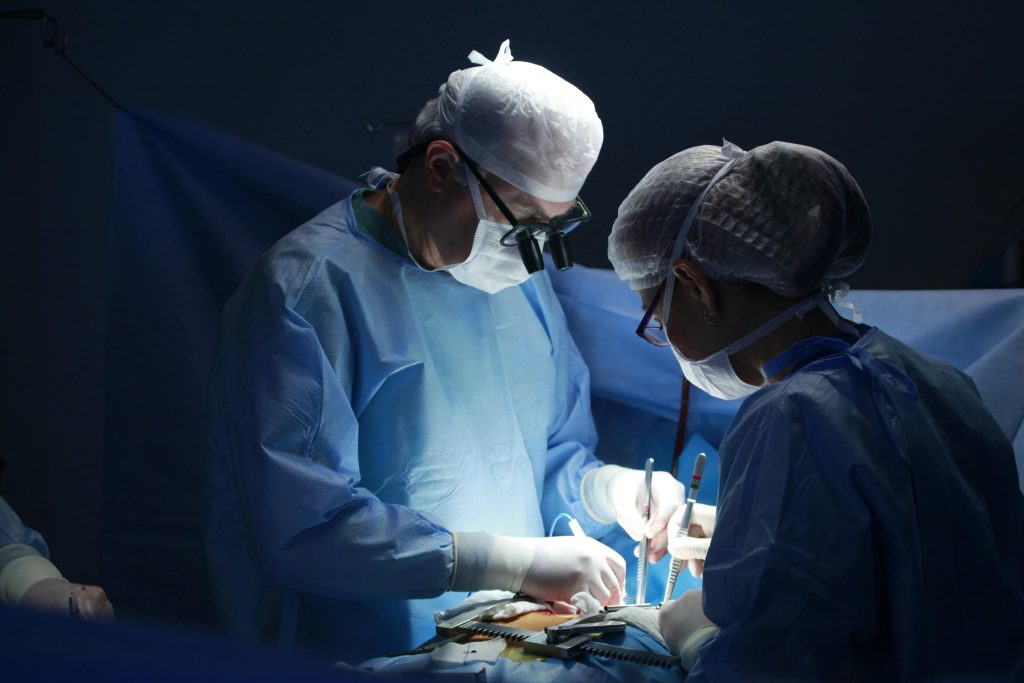Dois profissionais de saúde vestidos com roupa cirúrgica completa e máscaras, preparando um paciente masculino para uma operação numa sala de cirurgia. A cena mostra atenção à monitorização e aos cuidados pré-operatórios, com ambiente esterilizado e equipamentos médicos ao redor.