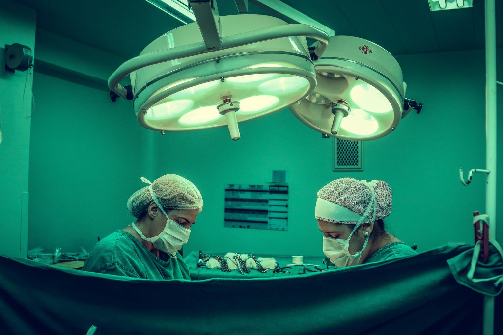 Dois profissionais de saúde vestidos com roupas cirúrgicas completas e luvas, realizando um procedimento dentro de uma sala de cirurgia. A mesa de instrumentos e o ambiente esterilizado de bloco operatório são visíveis, com foco na precisão e no trabalho em equipa durante o ato cirúrgico.