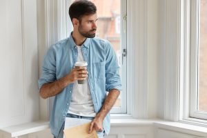 Homem adulto em pé junto a uma janela, segurando uma chávena de café, num momento de reflexão em ambiente interior com luz natural.