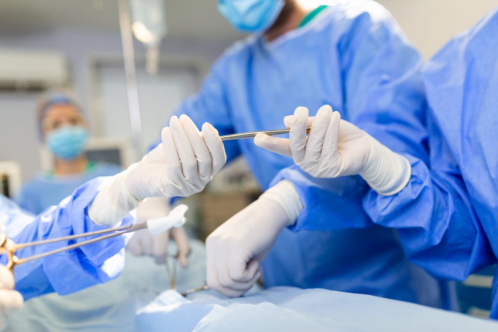 Fotografia de baixo ângulo numa sala de cirurgia, onde um assistente entrega instrumentos aos cirurgiões durante uma operação, mostrando equipa médica em ação.