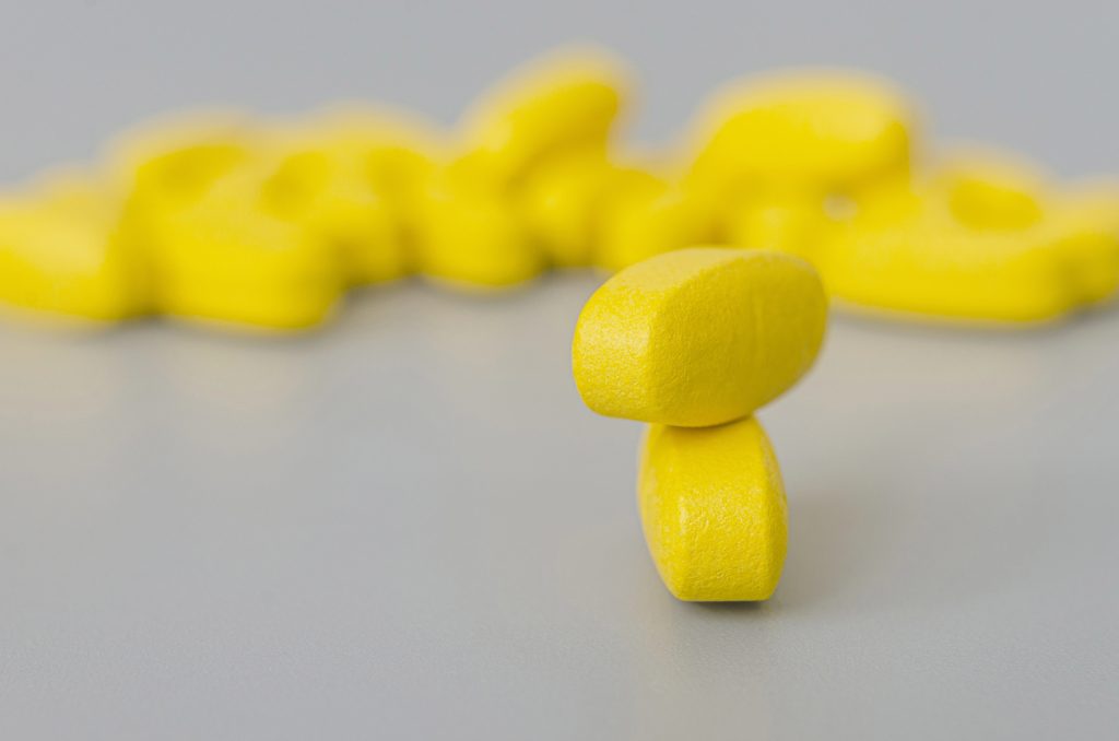 Dois comprimidos amarelos empilhados sobre um fundo acinzentado uniforme com pílulas semelhantes desfocadas atrás