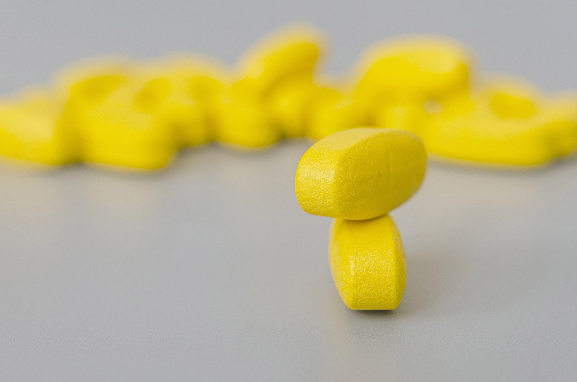 Dois comprimidos amarelos empilhados sobre um fundo acinzentado uniforme com pílulas semelhantes desfocadas atrás