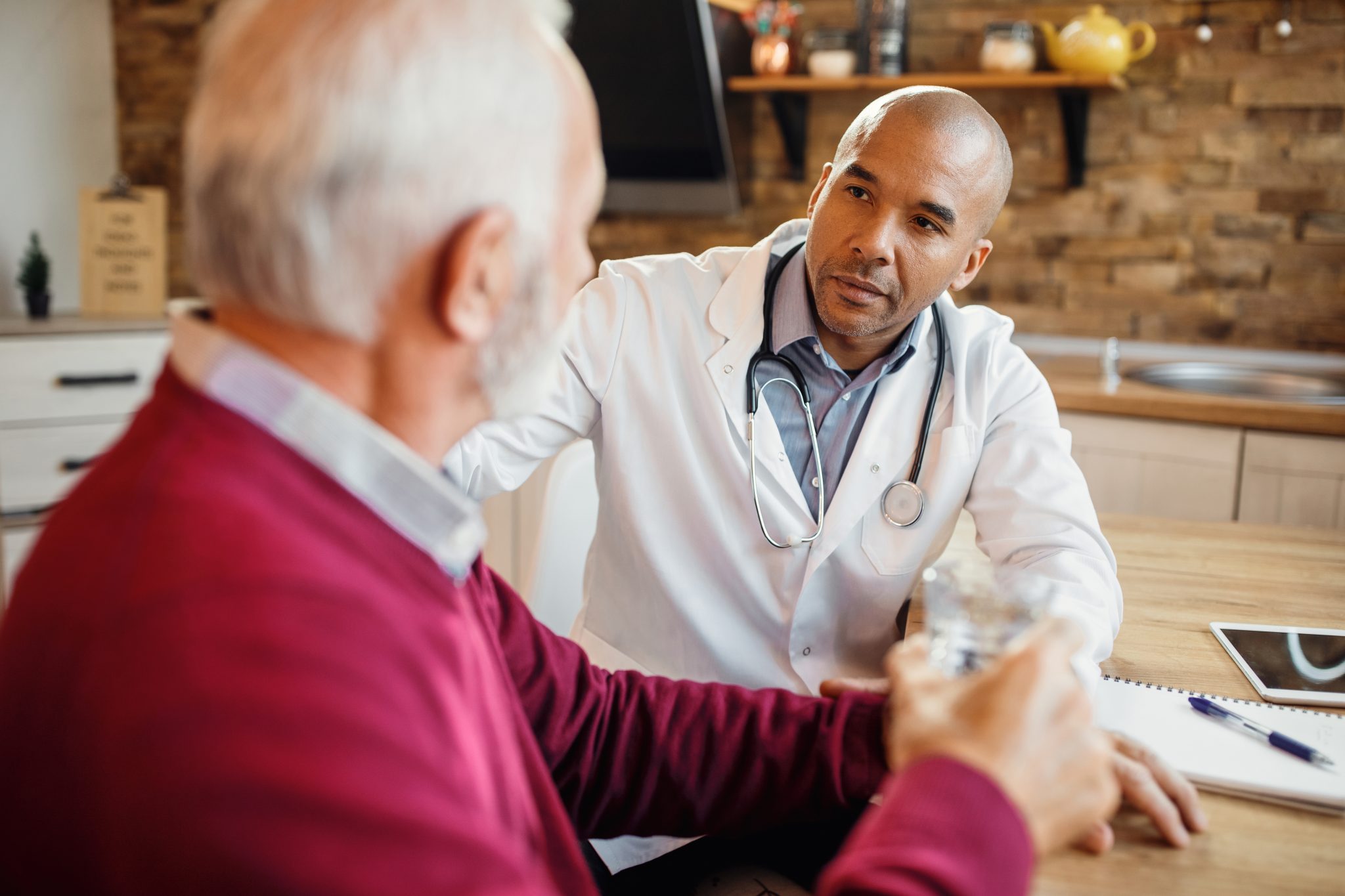 Alt texto: Médico a conversar com homem adulto durante consulta médica, sentado frente a frente num consultório, enquanto explica orientações de saúde.