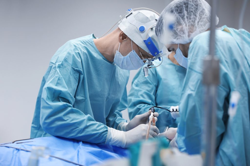 Surgeons wearing surgical clothing, leaning over a stretcher with a patient in a hospital setting during surgery for the treatment of Peyronie’s Disease.