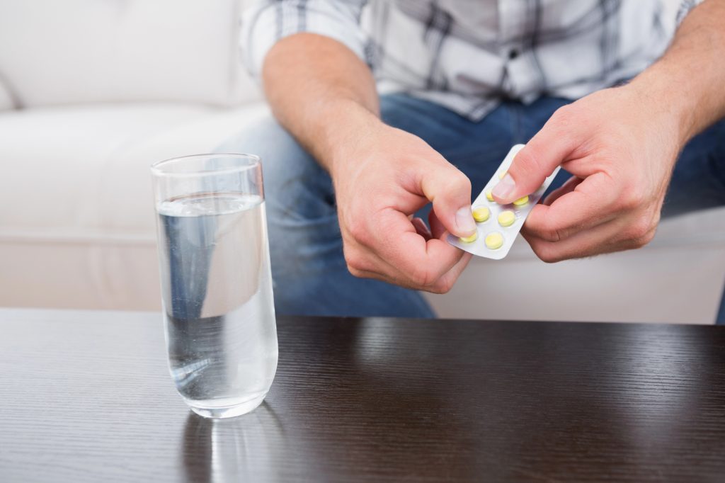 Homem de camisa aos quadrados sentado num sofá, com uma embalagem de Tadalafila 5 mg ou 20 mg na mão, e uma mesa de madeira com um copo de água.