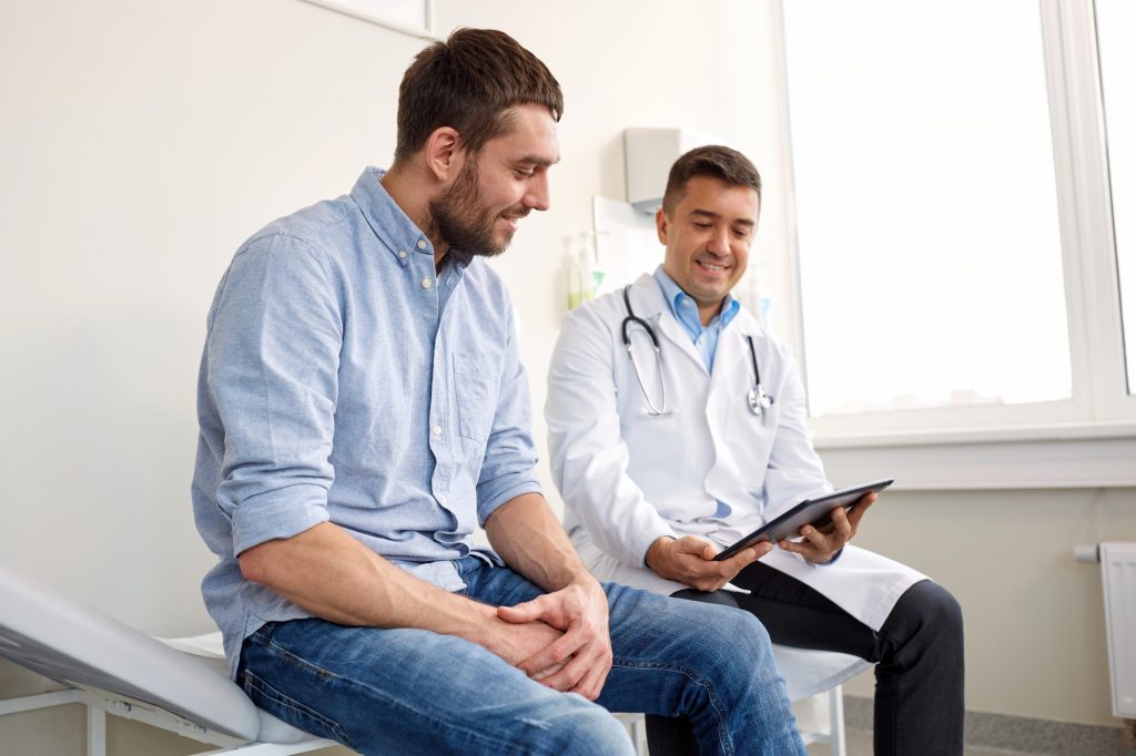 Médico urologista demonstra a um doente o funcionamento de uma prótese peniana insuflável através de um modelo gráfico durante uma consulta de acompanhamento pós-operatório.
