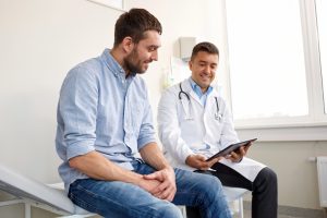 Médico urologista demonstra a um doente o funcionamento de uma prótese peniana insuflável através de um modelo gráfico durante uma consulta de acompanhamento pós-operatório.